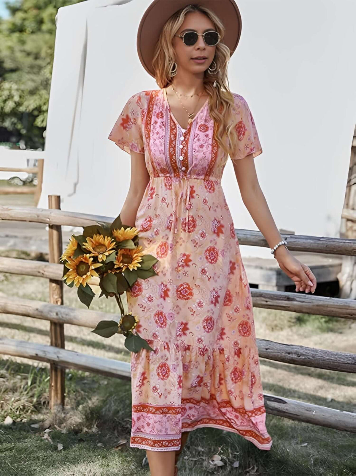 Woman in bohemian midi dress holding sunflowers, wearing sunglasses and a hat, against a rustic wooden fence.