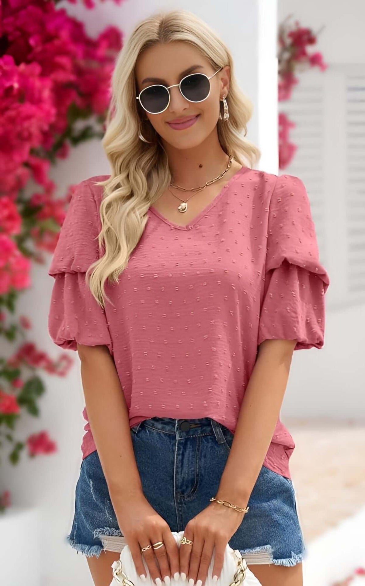 Woman wearing a pink puff sleeve blouse for women with Swiss dots, styled with denim shorts and sunglasses in a floral backdrop.